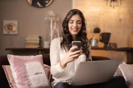 Mujer sonriendo mientras revisa su reembolso de impuestos FICA en su teléfono inteligente, sentada con su computadora portátil en casa, ilustrando la satisfacción de recuperar el dinero ganado con esfuerzo.