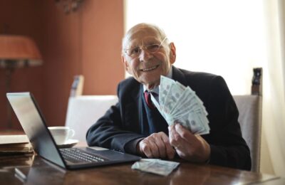 Senior businessman smiles while holding cash, demonstrating potential success from claiming Florida unclaimed property, with laptop nearby