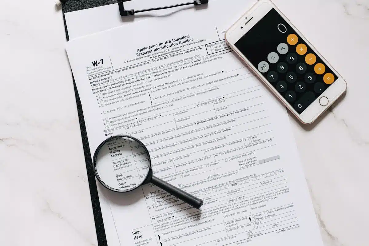 Form W-7 ITIN application on clipboard with magnifying glass and smartphone calculator, illustrating the process of applying for an Individual Taxpayer Identification Number