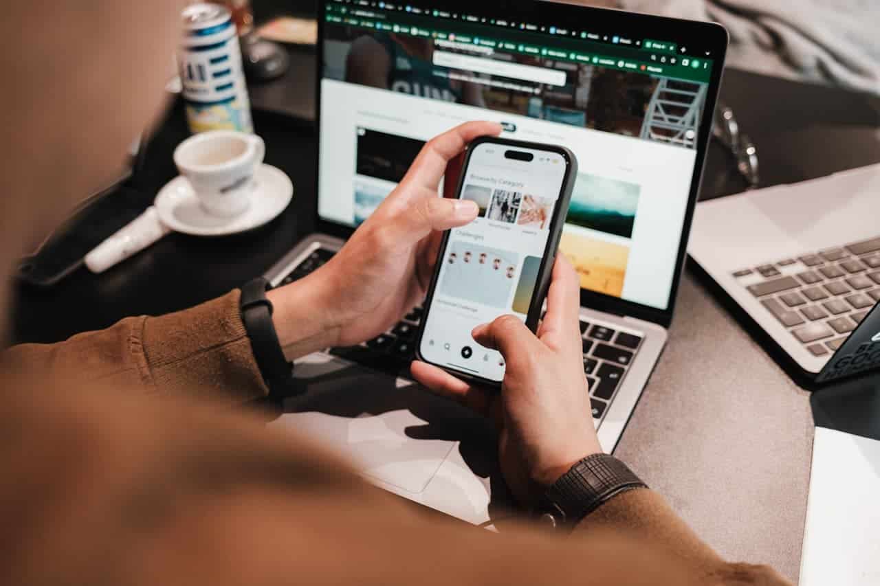 Person setting up an Id.me account on smartphone while working on laptop, with coffee and notepad nearby, illustrating secure digital identity verification process.