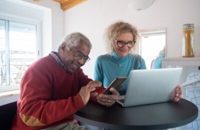 Couple de seniors utilisant un ordinateur portable et un smartphone pour faire des recherches et discuter de l'ajustement au coût de la vie (cola) de la sécurité sociale.