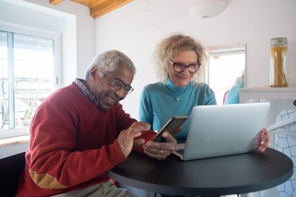 Pareja de personas mayores utilizando una computadora portátil y un teléfono inteligente para investigar y hablar sobre el ajuste por costo de vida (COLA) de la seguridad social.