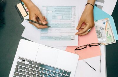 Hands filling out tax form with pen, surrounded by laptop, money, and glasses, illustrating the process of preparing taxes and checking Where's my refund status.