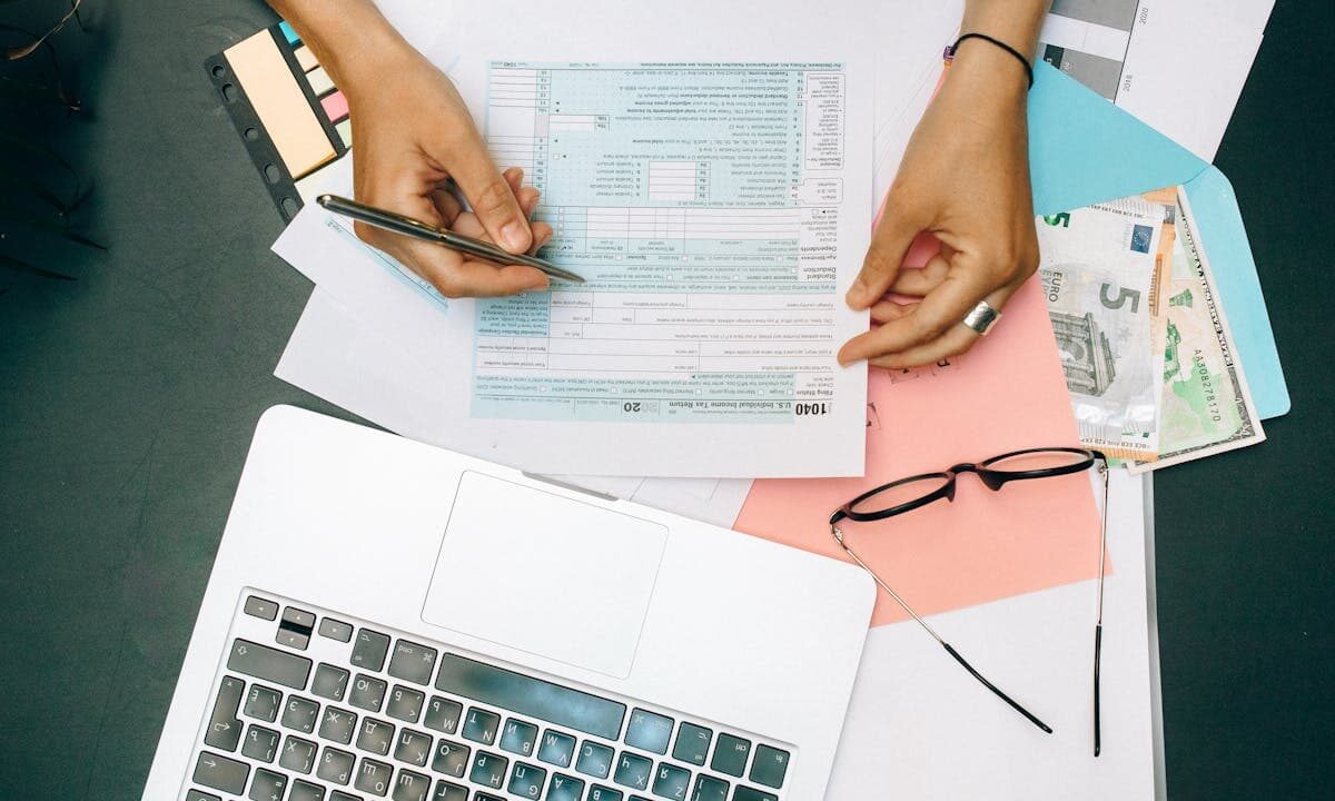 Hands filling out tax form with pen, surrounded by laptop, money, and glasses, illustrating the process of preparing taxes and checking Where's my refund status.