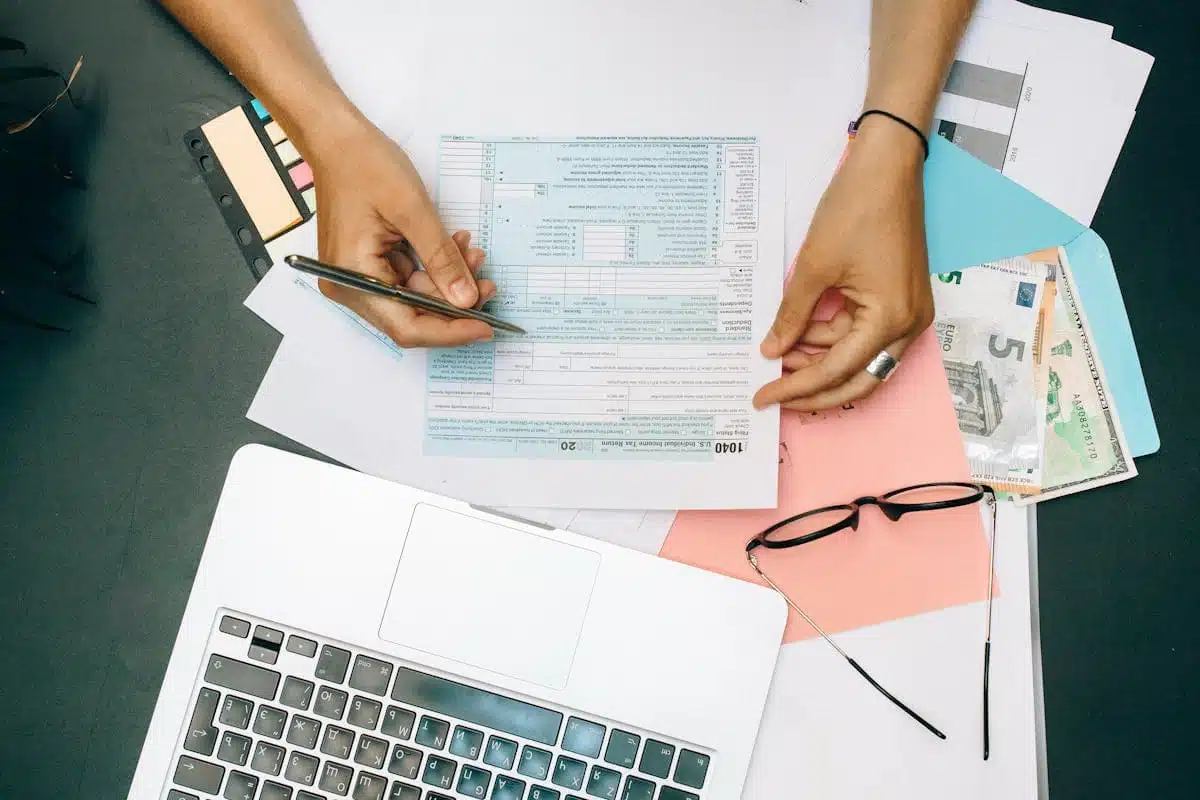 Hands filling out tax form with pen, surrounded by laptop, money, and glasses, illustrating the process of preparing taxes and checking Where's my refund status.