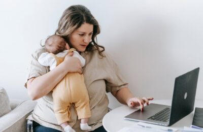 Multitasking mother holds baby while checking laptop, possibly seeing: your tax return is still being processed, balancing parenting and financial responsibilities.