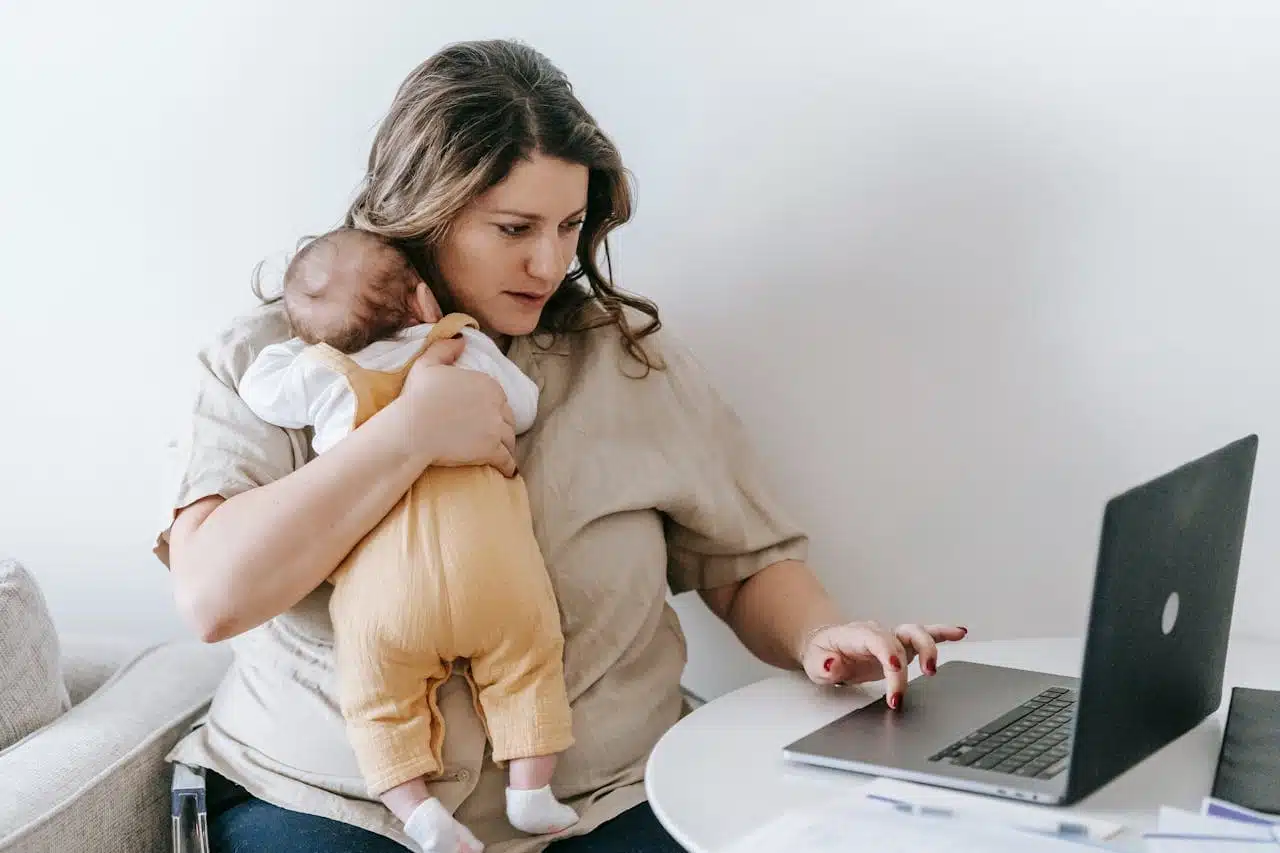 Multitasking mother holds baby while checking laptop, possibly seeing: your tax return is still being processed, balancing parenting and financial responsibilities.